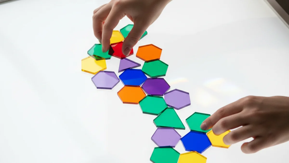 Mãos encaixando peças geométricas translúcidas coloridas sobre mesa iluminada, formando cascata modular.