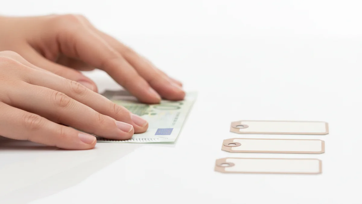 Mãos desdobrando uma cédula ao lado de etiquetas de preço em superfície branca.