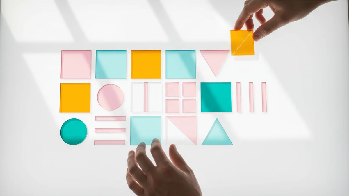 Mãos organizando peças geométricas coloridas translúcidas sobre mesa iluminada formando layout.